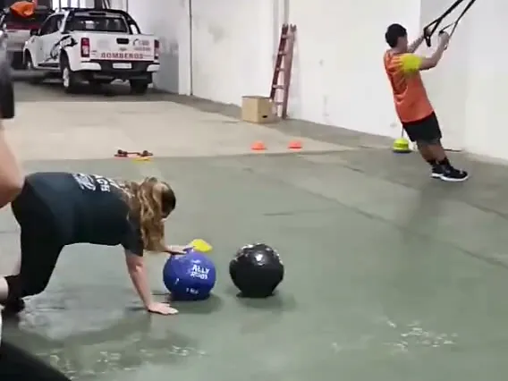 Una clase en el cuartel: un alumno hace remo con TRX contra la pared y una alumna, en cuatro apoyos, apoya una mano sobre una pelota medicinal. Atrás se ve una camioneta de Bomberos.